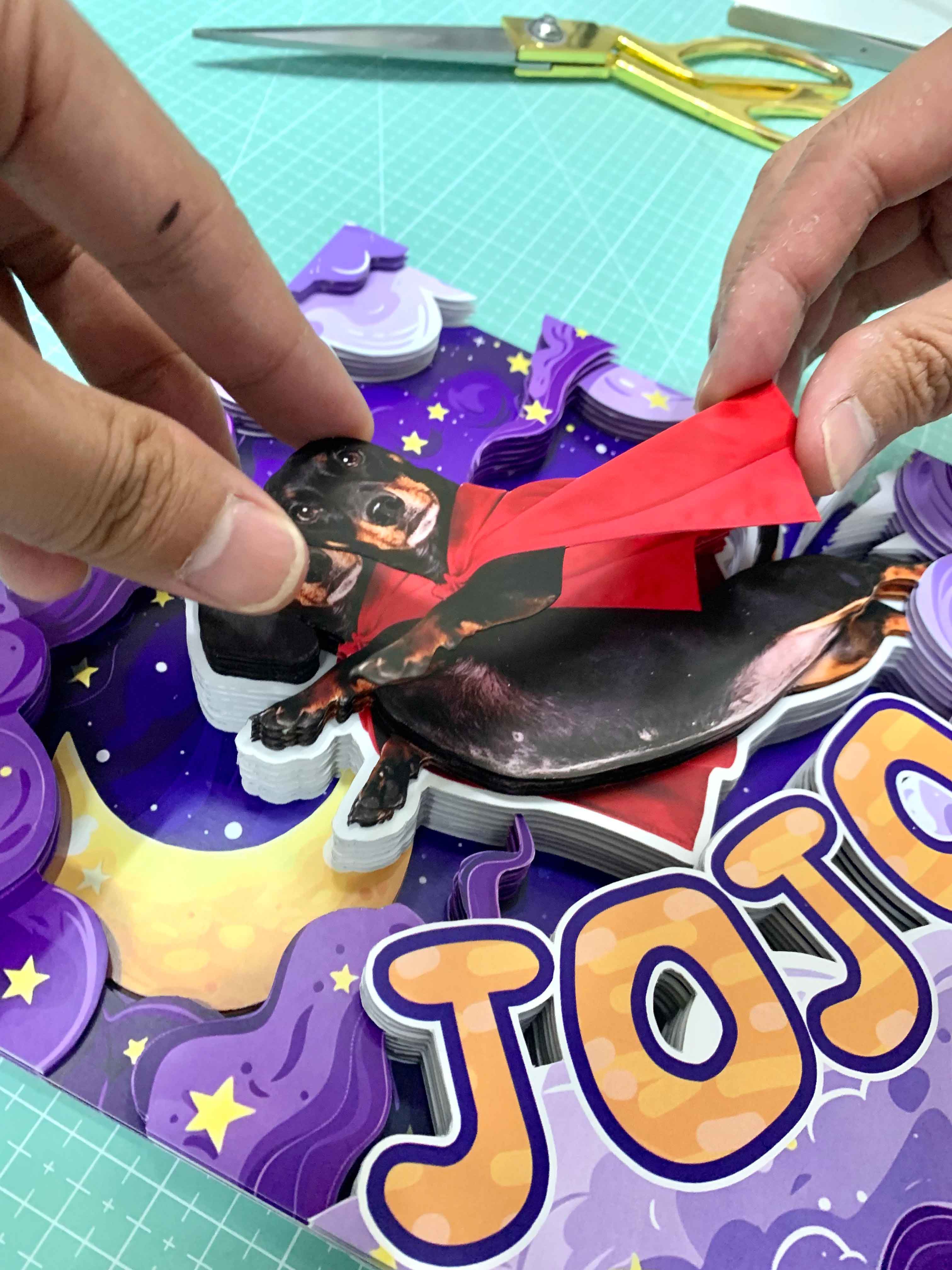 Artisan hands carefully placing a printed dachshund cutout wearing a red superhero cape onto a layered purple galaxy-themed custom 3D shadow box, with stacked paper pieces and gold scissors on a green cutting mat.